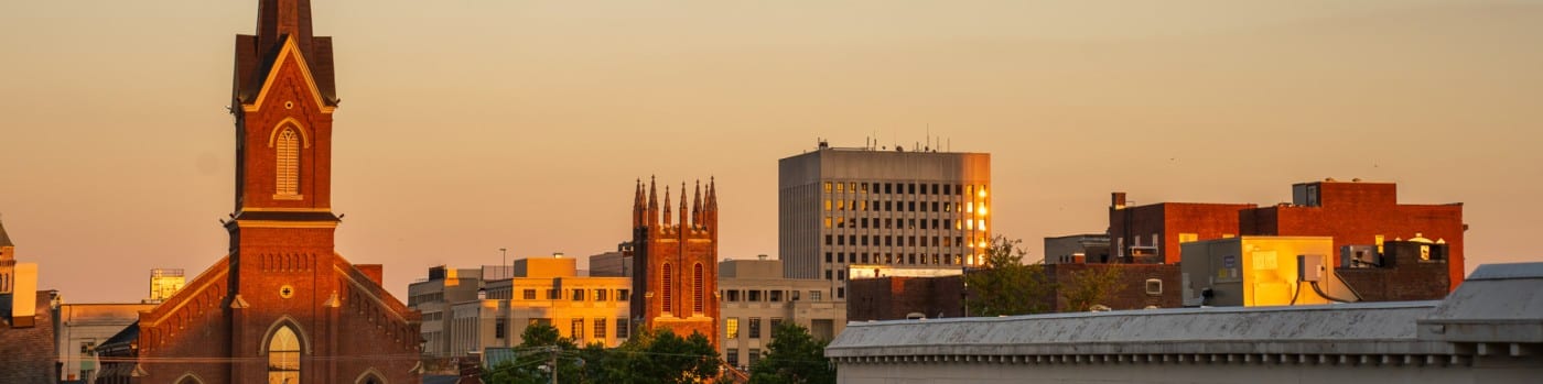 Downtown Lexington, Kentucky at golden hour: a red brick church steeple, a Gothic brick tower with pinnacles and office buildings glowing under a peach evening sky.